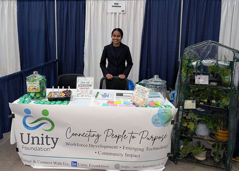 A Unity Foundation intern standing at the Unity Foundation booth, with a display greenhouse and Connecting People to Purpose banner, during the AFCEA TechNet Midwest conference.