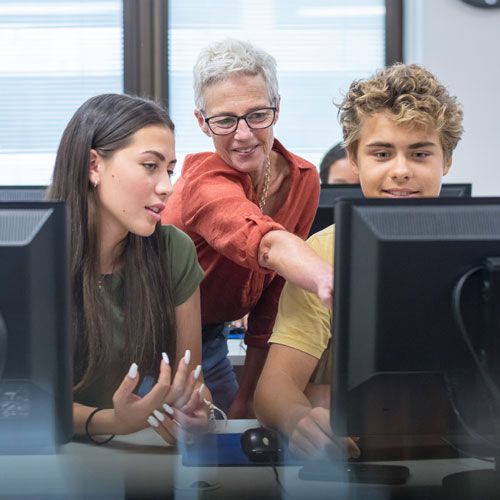 A mentor leans in to help two high-school students at their workstations.