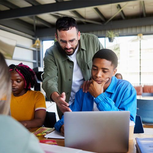 An engineer mentors a young student at a laptop while classmates collaborate around them.
