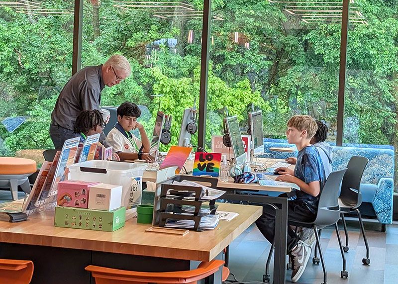 A Unity Foundation volunteer leaning in to help two campers at their workstations, with more campers working at screens along a window wall.