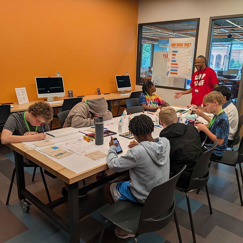 Campers gathered around a large table for a hands-on design activity while a camp staff member explains the session plan.